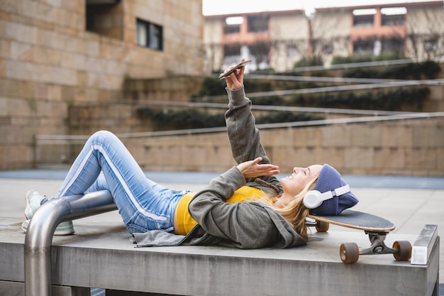 Mujer joven acostada con la cabeza en el skate sosteniendo un teléfono inteligente para hacer una llamada escuchando música y disfrutando de los contenidos en línea