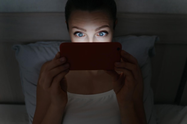 Mujer joven acostada en la cama y mirando adictivamente su teléfono inteligente a altas horas de la noche. Centrado en los ojos. Concepto de adicción a las redes sociales e intermet