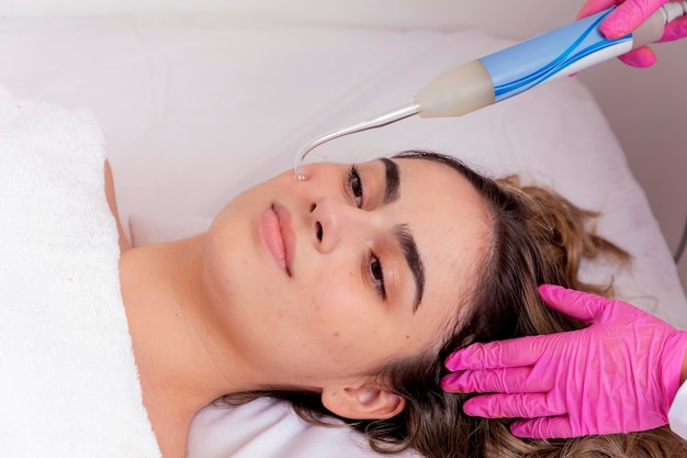 Mujer joven acostada en la mesa de la cosmetóloga durante el procedimiento de rejuvenecimiento La cosmetóloga cuida la piel del cuello y la cara joven y el bienestar Procedimiento de limpieza facial de hardware