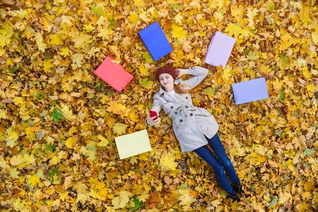 Mujer joven acostada sobre hojas de otoño y hace selfie rodeado de bolsas de la compra.