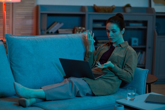 Mujer joven acostada en el sofá con el portátil en las piernas y hablando en línea con su amiga en la habitación de casa
