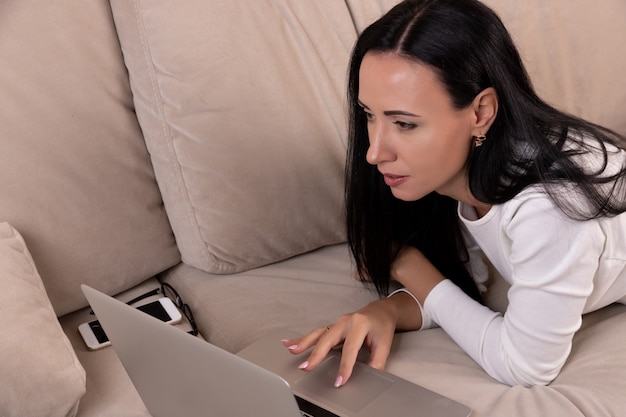 Mujer joven acostada en un sofá y usa una computadora portátil