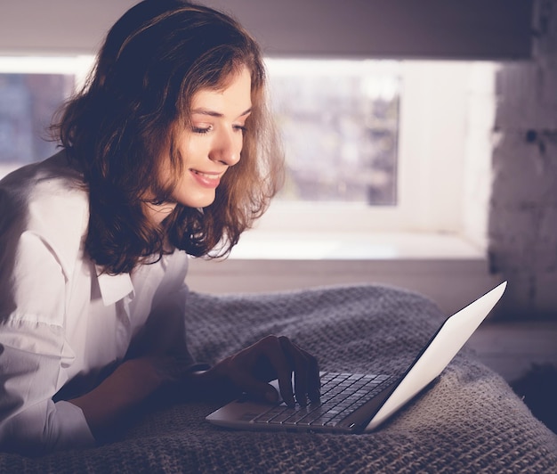 Mujer joven se acuesta en la cama con una computadora portátil Concepto de estilo de vida y personas