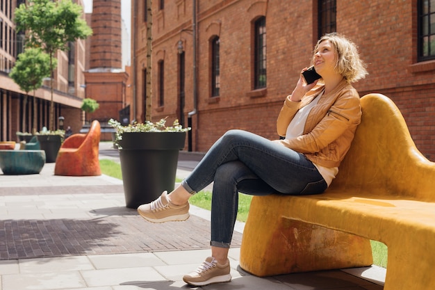 Mujer joven admira la arquitectura urbana mientras está sentada en un banco en la ciudad y habla por teléfono. Comunicación móvil. Turismo y tecnologías modernas. Recreación y entretenimiento Vida moderna.