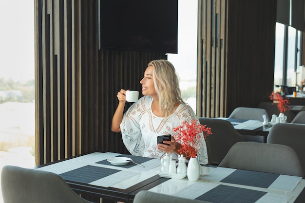 Mujer joven adulta hermosa y feliz rubia en un restaurante para desayunar con un teléfono inteligente