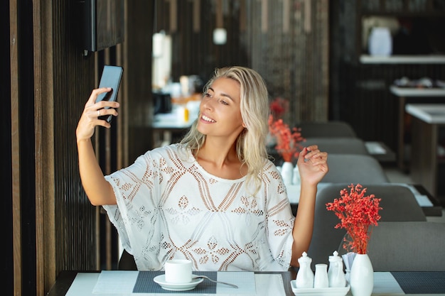 Mujer joven adulta hermosa y feliz rubia en un restaurante para desayunar con un teléfono inteligente