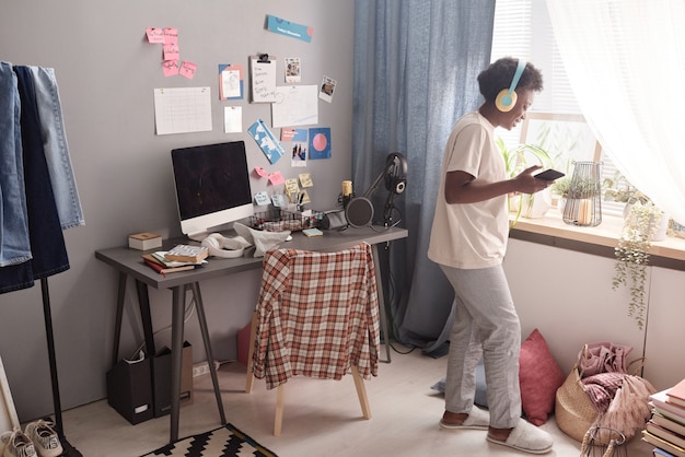 Mujer joven africana en auriculares inalámbricos con su teléfono móvil para escuchar música en la habitación