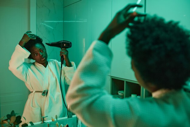 Mujer joven africana en bata de baño mirando en el espejo y secándose el cabello con secador de pelo en el baño