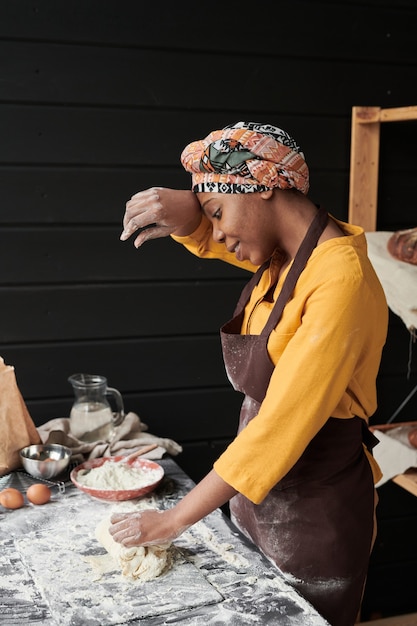 Mujer joven africana en delantal de pie en la mesa y amasando la masa en la cocina