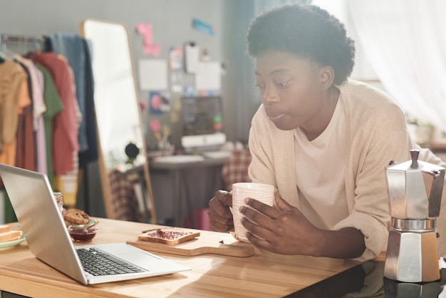 Mujer joven africana desayunando con tostadas y mermelada y viendo algo en la computadora portátil mientras está sentado en la mesa de su habitación