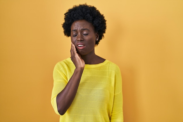Mujer joven africana de pie sobre un estudio amarillo tocando la boca con la mano con expresión dolorosa debido a dolor de muelas o enfermedad dental en el dentista dental