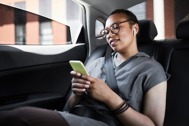 Mujer joven africana sentada en el asiento trasero y leyendo un mensaje en el teléfono móvil durante su viaje en el coche