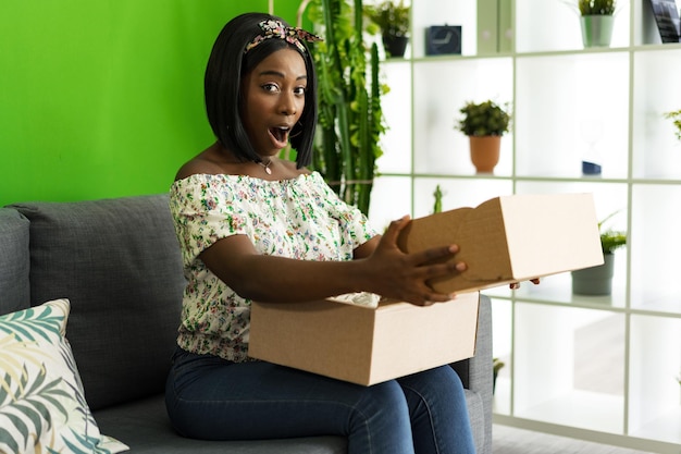 Mujer joven africana sentada en el sofá en casa desempacando caja de cartón con compra en línea