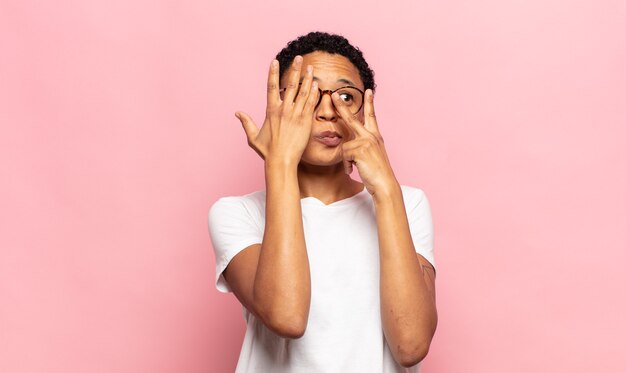 Mujer joven afro que cubre la cara con las manos, mirando entre los dedos con expresión de sorpresa y mirando hacia un lado