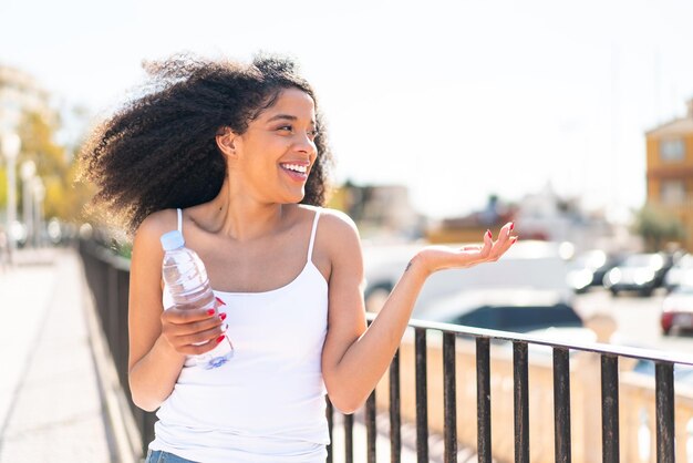 Mujer joven afroamericana con una botella de agua al aire libre con una expresión facial de sorpresa