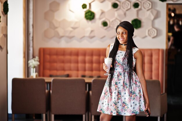 Mujer joven afroamericana pura en vestido de verano y collar de gargantilla en el café bebiendo cóctel de batido