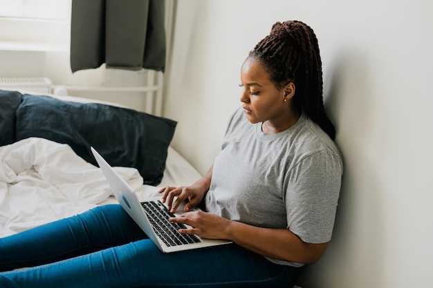 Mujer joven afroamericana que usa una computadora portátil en las tecnologías de la cama y la comunicación y la red social