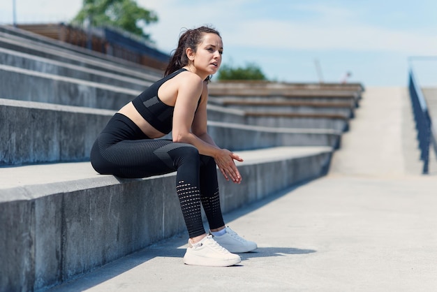 Mujer joven agotada después del entrenamiento sentado en las escaleras en el estadio deportivo