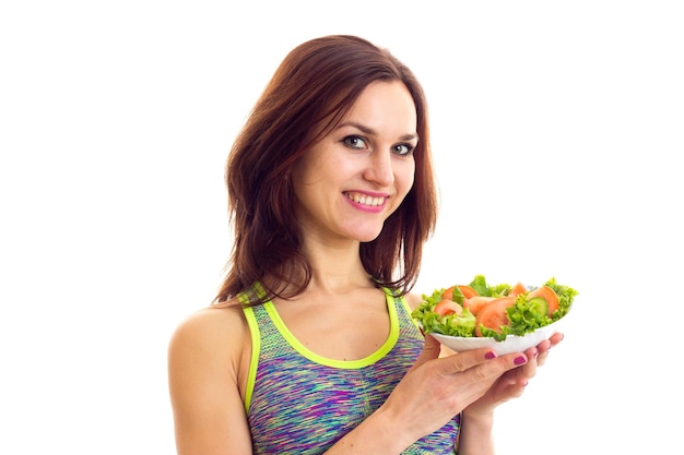 Una mujer joven y agradable vestida con una camiseta deportiva de colores sosteniendo un tenedor y un plato con ensalada verde y tomate