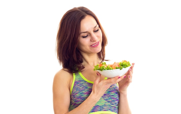 Una mujer joven y agradable vestida con una camiseta deportiva de colores sosteniendo un tenedor y un plato con ensalada verde y tomate