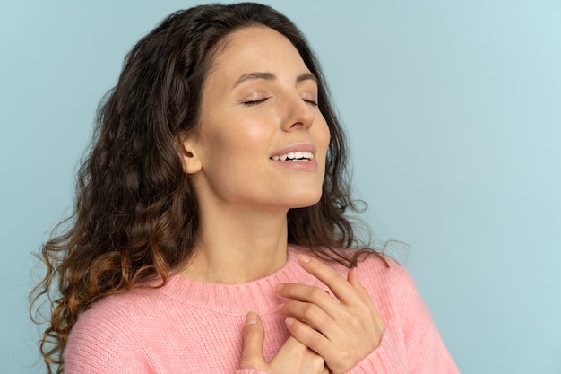 Mujer joven agradecida con los ojos cerrados sonriendo agradeciendo a Dios, respira aire fresco, muestra fe y amor.