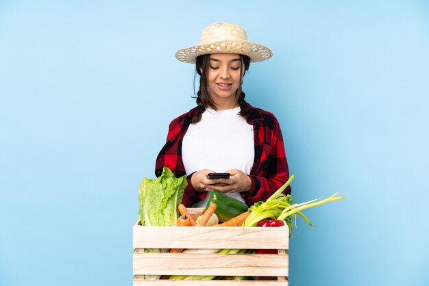 Mujer joven agricultor sosteniendo verduras frescas en una canasta de madera enviando un mensaje con el móvil