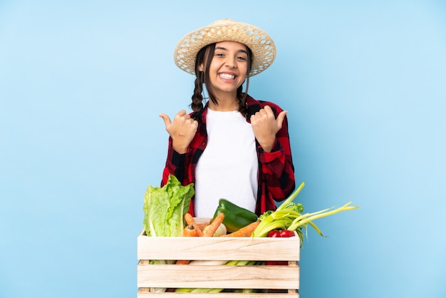 Mujer joven agricultor sosteniendo verduras frescas en una canasta de madera con gesto de pulgar hacia arriba y sonriendo