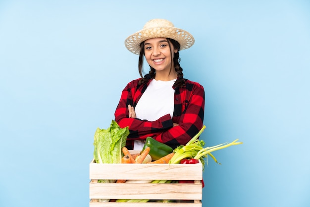 Mujer joven agricultor sosteniendo verduras frescas en una canasta de madera manteniendo los brazos cruzados en posición frontal