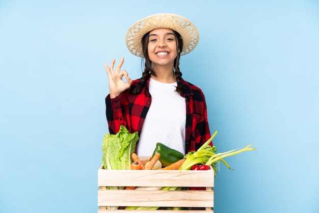 Mujer joven agricultor sosteniendo verduras frescas en una canasta de madera mostrando signo ok con los dedos