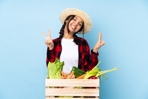 Mujer joven agricultor sosteniendo verduras frescas en una canasta de madera que muestra el signo de la victoria con ambas manos