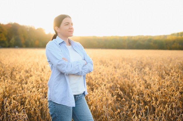 Mujer joven agricultora de pie en el campo de soja en verano sosteniendo una tableta y monitoreando el crecimiento de los cultivos