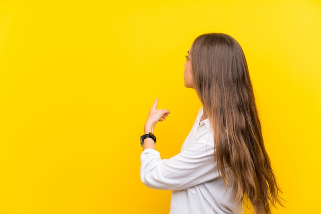 Mujer joven aislada en amarillo apuntando hacia atrás con el dedo índice