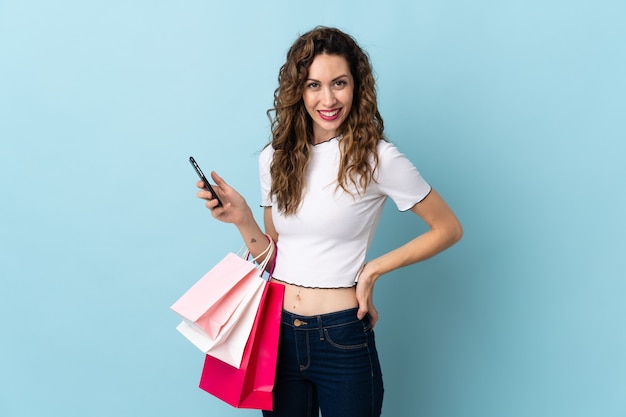 Mujer joven aislada en la pared azul sosteniendo bolsas de la compra y escribiendo un mensaje con su teléfono celular a un amigo