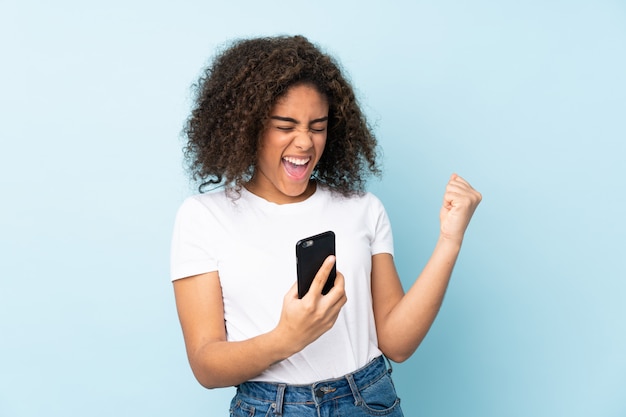 Mujer joven aislada en la pared azul con teléfono en posición de victoria