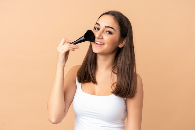 Mujer joven aislada en la pared beige con pincel de maquillaje y con expresión feliz