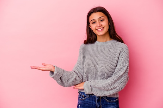 Mujer joven aislada en la pared rosa mostrando un espacio de copia en una palma y sosteniendo otra mano en la cintura