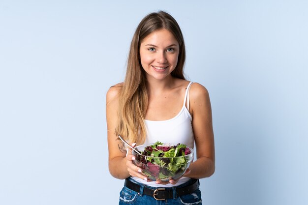 Mujer joven aislada sobre fondo azul sosteniendo un plato de ensalada con expresión feliz