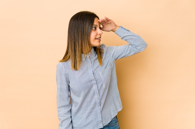 Mujer joven aislada sobre fondo beige mirando lejos manteniendo la mano en la frente.