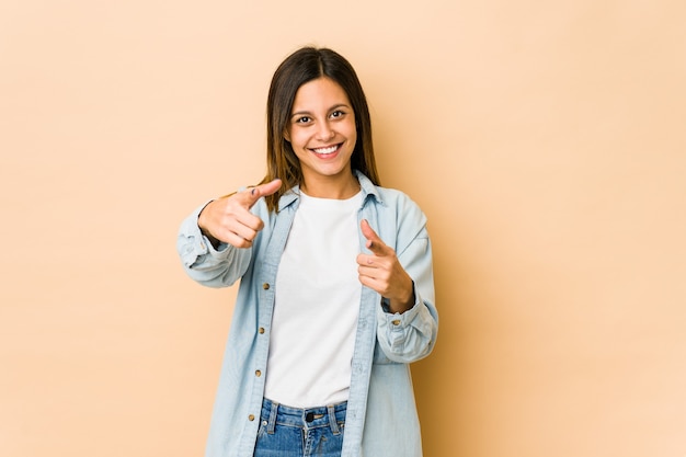 Mujer joven aislada sobre fondo beige sonrisas alegres apuntando al frente.