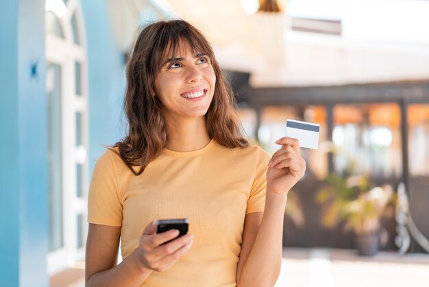 Mujer joven al aire libre comprando con el móvil con tarjeta de crédito mientras piensa