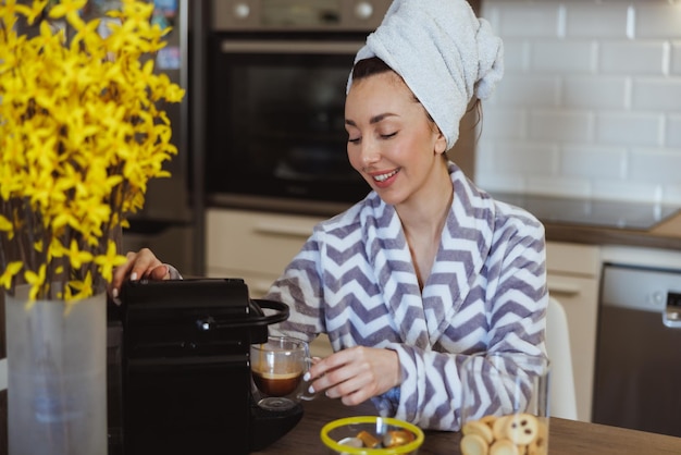 Mujer joven en albornoz haciendo café con una máquina de café en la cocina de casa.