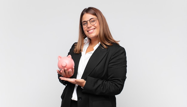 Mujer joven con una alcancía sonriendo alegremente, sintiéndose feliz y mostrando un concepto en el espacio de la copia con la palma de la mano