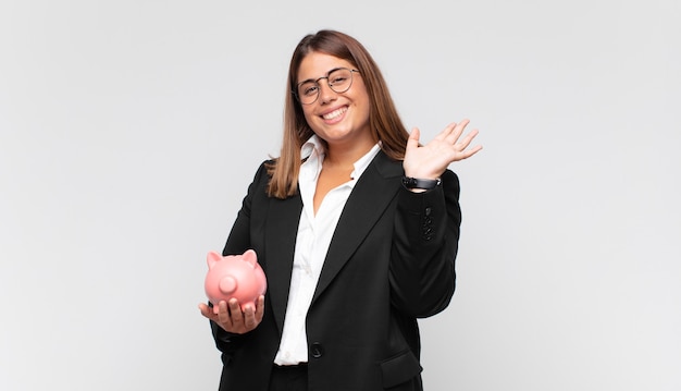 Mujer joven con una alcancía sonriendo feliz y alegremente, saludando con la mano, dándote la bienvenida y saludándote, o despidiéndote