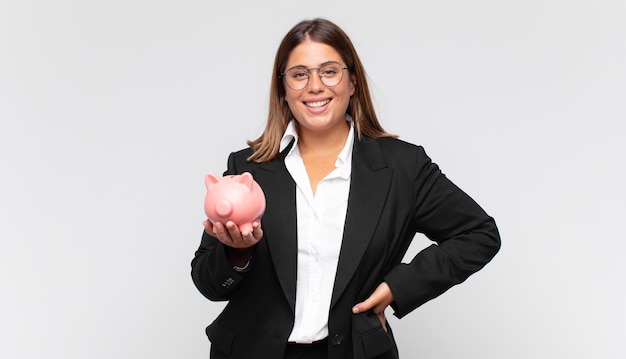 Mujer joven con una alcancía sonriendo felizmente con una mano en la cadera y una actitud segura, positiva, orgullosa y amistosa