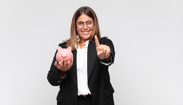 Mujer joven con una alcancía sonriendo con orgullo y confianza haciendo la pose número uno triunfalmente, sintiéndose como una líder