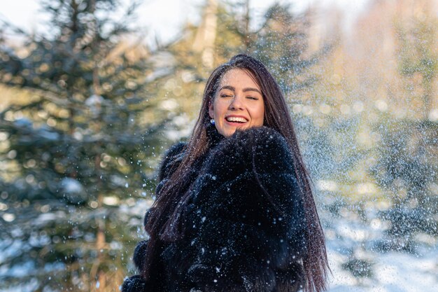 Mujer joven alegre en un abrigo de piel caliente disfrutando de un día de invierno en el bosque nevado