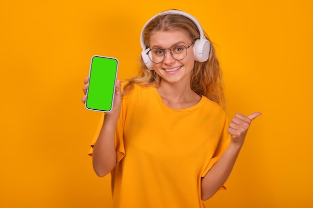 Mujer joven y alegre adolescente caucásica con auriculares mostrando el teléfono y el pulgar hacia arriba