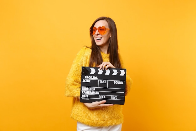 Mujer joven alegre en anteojos de corazón naranja suéter mirando a un lado sosteniendo claqueta de cine negro clásico aislado sobre fondo amarillo. Estilo de vida de las emociones sinceras de la gente. Área de publicidad.