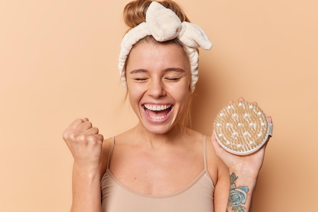 La mujer joven alegre aprieta el puño con expresión feliz se regocija de éxito sostiene cepillo de baño lleva diadema y camiseta aislada sobre fondo marrón. Rutina matutina y concepto de tratamiento corporal.