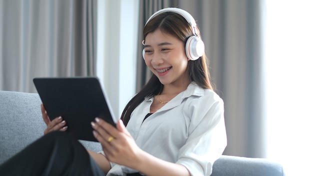 Una mujer joven alegre con auriculares está usando tableta para ver películas en línea en el sofá de la sala de estar.